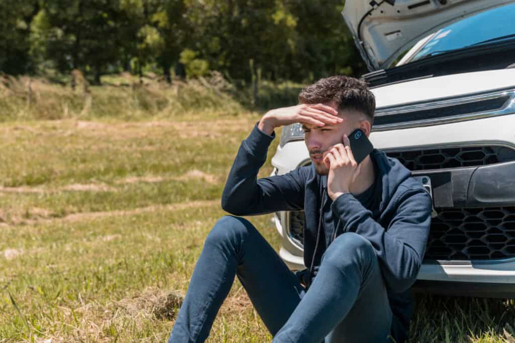 A concerned man sitting on the ground next to a car with its hood open, holding a phone to his ear and shielding his eyes from the sun.