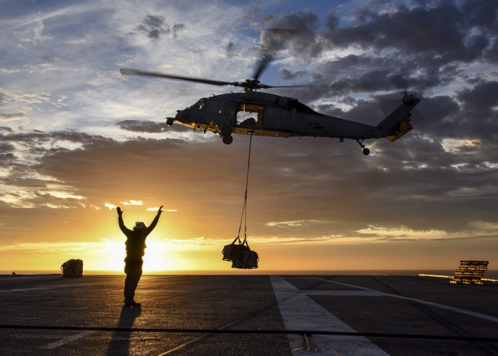 a man standing on a helipad while a cargo plane lifts goods upon takeoff