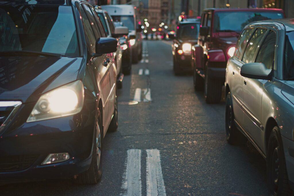 Cars stuck in traffic on a city street in the early evening with headlights on.