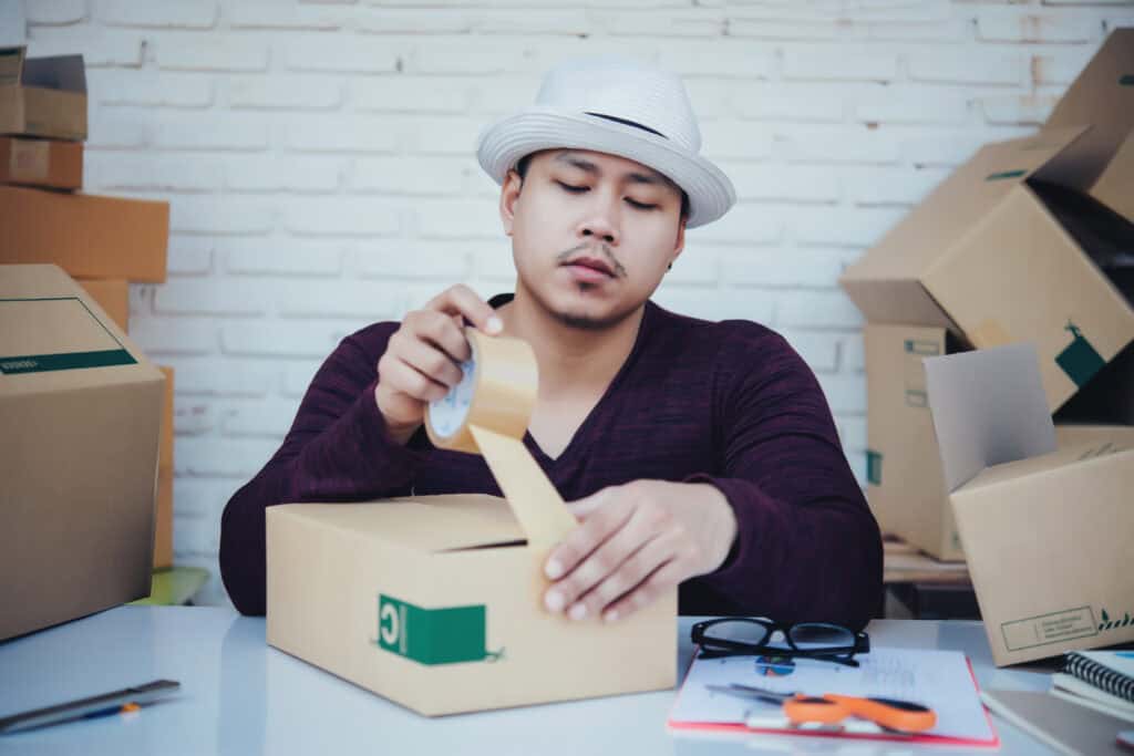 A man in a hat carefully sealing a cardboard box with packing tape, surrounded by packages, indicating the process of preparing an item for return or shipment.