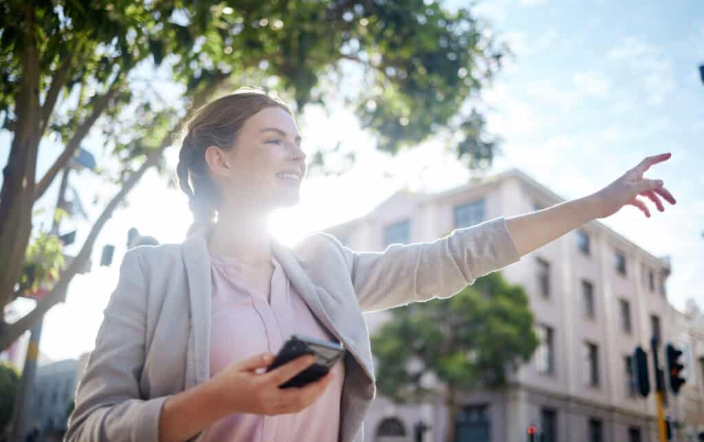 Mujer levanta el brazo en la calle contra sol mientras tiene el teléfono en la mano con la aplicación de Uber,
