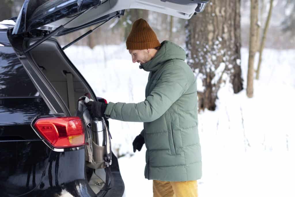 Un hombre con un abrigo de invierno verde y gorro toma una bolsa del maletero de su SUV en un bosque nevado.