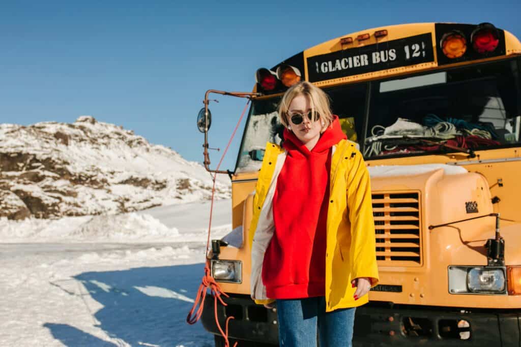 Persona con hoodie rojo y chaqueta amarilla de pie frente a un autobús escolar cubierto de nieve en condiciones invernales.