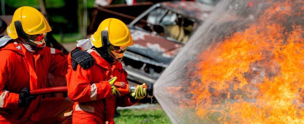 Bomberos combatiendo un incendio forestal con vehículos quemados al fondo